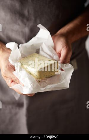 Mains d'un chef tenant un pat de la ferme frais beurre assaisonné avec des herbes dans un emballage ouvert de près vers le haut avec espace de copie vertical Banque D'Images
