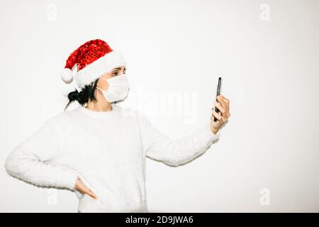 Une jeune femme avec un masque de visage tient un téléphone portable haut isolé sur fond blanc. Concept de facetime et de la communication pendant les vacances en cas de pandémie. Banque D'Images