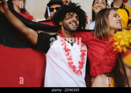 Couple avec guirlande portant un drapeau allemand dans la zone des fans du stade regardant un match. Groupe de jeunes hommes et femmes qui applaudissent l'équipe de football de l'allemagne. Banque D'Images