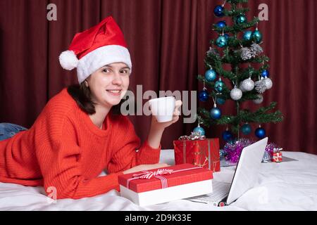 Une jeune femme dans un chapeau de père Noël et un chandail rouge tient une tasse de café ou de thé, repose avec son ordinateur portable, allongé sur le lit, entouré de cadeaux de Noël. C Banque D'Images