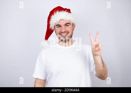 Jeune homme beau portant un chapeau de père Noël sur fond blanc afficher et pointer vers le haut avec les doigts numéro deux tout en souriant confiant et heureux Banque D'Images