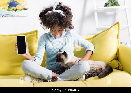 Fille afro-américaine souriante montrant un smartphone avec écran vierge, regardant le chat et assis sur un canapé à la maison Banque D'Images