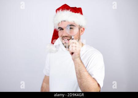 Jeune homme beau portant un chapeau de père Noël sur fond blanc surpris de regarder à travers une loupe Banque D'Images