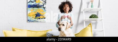 KIEV, UKRAINE - 02 OCTOBRE 2020: Happy girl assis derrière le labrador sur un canapé, tout en jouant avec joystick à la maison, bannière Banque D'Images