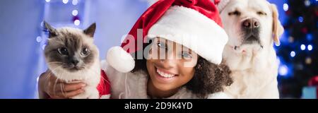 joyeux afro-américain fille dans chapeau de santa regardant l'appareil photo près de peluche chat et labrador chien sur fond flou, bannière Banque D'Images