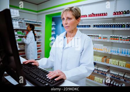 Pharmacienne caucasienne tapant sur un ordinateur de bureau en recherchant par e-mails debout en pharmacie Banque D'Images