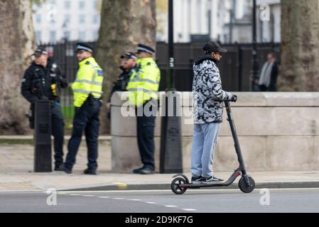 Jeune homme à bord d'un scooter électrique passé des policiers à Whitehall, Westminster, Londres, Royaume-Uni, Eco transport personnel illégalement monté sur la route Banque D'Images