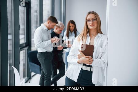 Portrait de blond gai avec bloc-notes qui se tient devant de jeunes succès équipe qui travaillent et communiquent ensemble à l'intérieur au bureau Banque D'Images