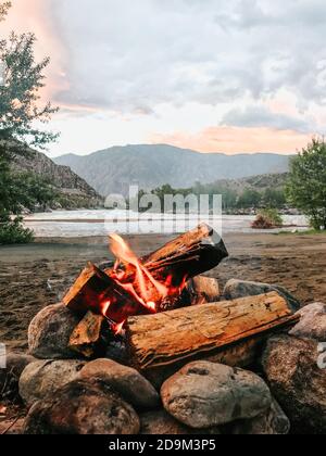 Feu de joie dans un camp dans les montagnes sur les rives d'une rivière rapide sur une soirée d'été au coucher du soleil Banque D'Images