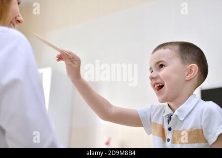 L'enfant souriant gaiement tire sa main avec une spatule au médecin en uniforme blanc. Banque D'Images