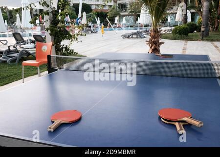 Belek, Turquie - octobre 2020 : tennis de table prêt pour les touristes à l'extérieur. Quatre raquettes sur une table bleue, une piscine et des chaises longues avec des voyageurs en arrière-plan. Banque D'Images