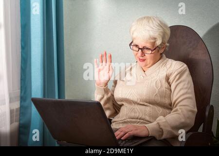 Une vieille dame s'assoit à la maison dans une chaise avec son ordinateur portable, communiquant en ligne, agitant sa main en bienvenue Banque D'Images