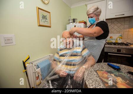 Homme âgé dans les années quatre-vingt ayant ses cheveux coupés par un coiffeur portant un EPI pour la protection contre le coronavirus tout en travaillant à distance, Angleterre, Royaume-Uni Banque D'Images