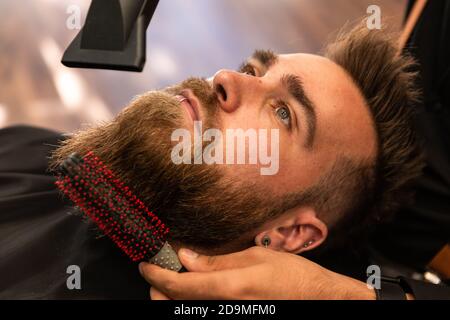 Un garçon caucasien avec une barbe est peigné par un coiffeur hispanique dans le salon de coiffure avec un peigne et un sèche-cheveux. Zenithal avion. Banque D'Images