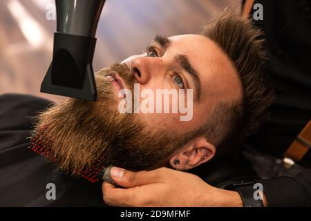 Un garçon caucasien avec une barbe est peigné par un coiffeur hispanique dans le salon de coiffure avec un peigne et un sèche-cheveux. Zenithal avion. Banque D'Images