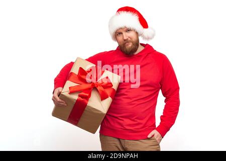 Un homme brutal avec une barbe rouge dans un chapeau de père noël regarde la caméra et tient une boîte cadeau avec un ruban rouge dans ses mains. Studio shot Isolate, chris Banque D'Images