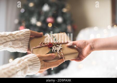 Femme en chandail donnant une boîte cadeau de Noël emballée à l'enfant. Bokeh de neige brillant, sapin. Vacances d'hiver. Banque D'Images