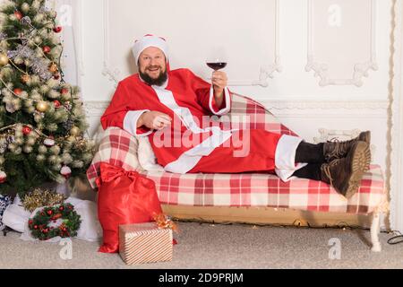 Homme barbu vêtu du costume du Père Noël assis sur un canapé avec un verre de vin portrait de studio Banque D'Images