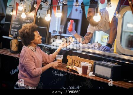 jeune employé de beardy dans la restauration rapide ajoutant une ketchup un sandwich à une cliente afro-américaine satisfaite Banque D'Images