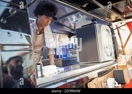 jeune afro-américaine employée dans le service de restauration rapide écrivant la commande regardant le jeune race blanche beardy hipster avec une casquette. Réflexion. Banque D'Images