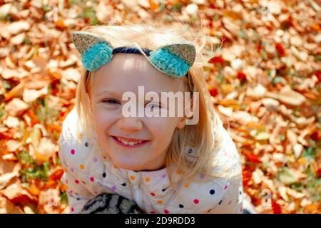 Adorable fille blonde assise sur des feuilles jaunes déchue regardant vers le haut un beau jour d'automne. Jouer à l'extérieur dans le parc. Fille portant un serre-tête de chat. Banque D'Images