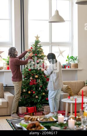 Couple mature contemporain en vêtements décontractés décorant arbre de Noël avec jouets Banque D'Images