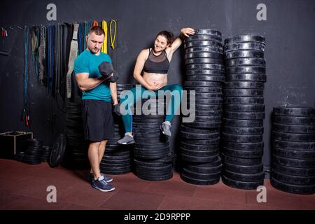 Athlètes femme enceinte et son mari posant dans la salle de gym près de l'équipement sportif. Grossesse sportive active. Faites de l'exercice dans la salle de sport pendant Banque D'Images