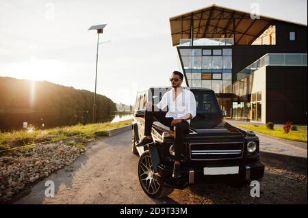 Homme asiatique solide en chemise blanche et lunettes de soleil posé près de la voiture suv mafia noire. Banque D'Images