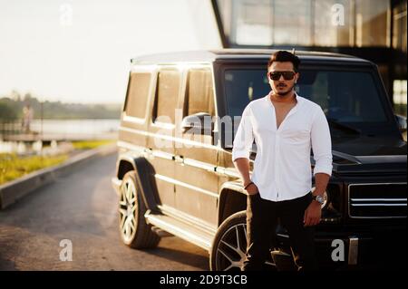 Homme asiatique solide en chemise blanche et lunettes de soleil posé près de la voiture suv mafia noire. Banque D'Images