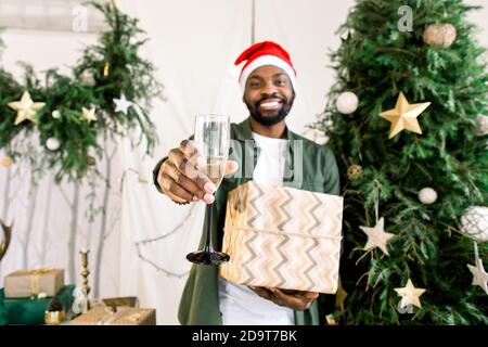 Souriant homme africain attrayant dans chapeau de Santa près de l'arbre de Noël tenant le champagne et le cadeau . Lumières Bengale. Chambre confortable. Noël, nouvel an, hiver Banque D'Images