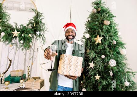 Souriant homme africain attrayant dans chapeau de Santa près de l'arbre de Noël tenant le champagne et le cadeau . Lumières Bengale. Chambre confortable. Noël, nouvel an, hiver Banque D'Images