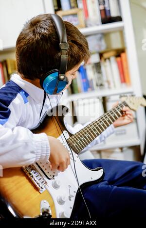 enfant jouant de la guitare électrique à la maison avec une paire d'écouteurs et une bibliothèque non focalisée en arrière-plan, mise au point sélective, cadrage vertical Banque D'Images