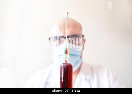 gros plan d'une seringue de sang avec une goutte sur le pointe de l'aiguille avec le médecin hors foyer en arrière-plan, mise au point sélective Banque D'Images