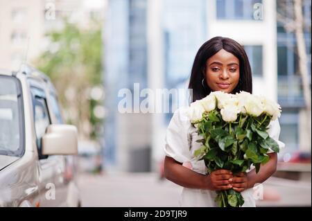 Belle fille afro-américaine tenant bouquet de fleurs roses blanches sur la datation dans la ville. Femme d'affaires noire avec bouquet de fleurs près de la voiture. Banque D'Images