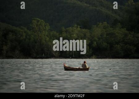 Un pêcheur de subsistance travaillant sur un petit bateau en bois, flottant sur la baie de Tapian Nauli au large des rives de la ville de Sibolga avec une zone de forêt de plaines peut être vu en arrière-plan. Province de Sumatra Nord, Indonésie. Banque D'Images