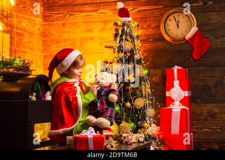 Garçon en costume de père Noël tenant un ours en peluche et attendant New Yeear minuit. Magnifique arbre décoré de Noël. Cadeaux et cadeaux de vacances d'hiver. Heureux Banque D'Images