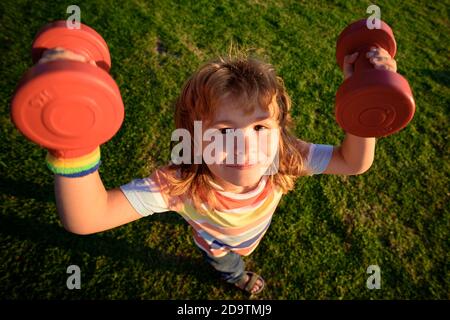 Sport amusant pour enfants. Enfant avec haltères dans le parc. Petit garçon fort. Visage d'enfant amusant. Développement des enfants et exercice sain et fort. Grand angle. Banque D'Images