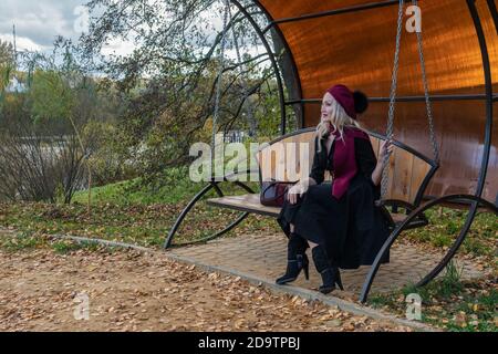 Une jeune fille souriante est assise sur une balançoire de jardin, forgée dans un manteau bordeaux et le biret, avec un beau sourire, à l'automne contre le fond des arbres, le Banque D'Images