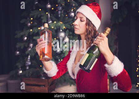 Une jeune femme en costume du Père Noël tient une bouteille de champagne et de whisky. Choisir une boisson alcoolisée pour les vacances. Banque D'Images