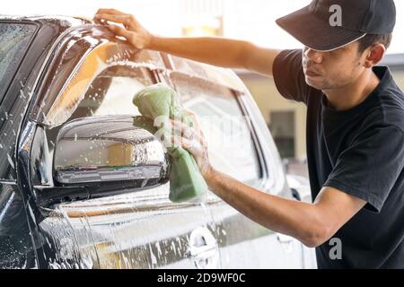 Le travailleur de lavage de voiture portant un T-shirt et un capuchon noir utilise une éponge pour nettoyer la voiture dans le centre de lavage de voiture, concept pour l'industrie de l'entretien de voiture. Banque D'Images