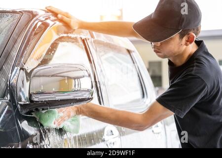 Le travailleur de lavage de voiture portant un T-shirt et un capuchon noir utilise une éponge pour nettoyer la voiture dans le centre de lavage de voiture, concept pour l'industrie de l'entretien de voiture. Banque D'Images