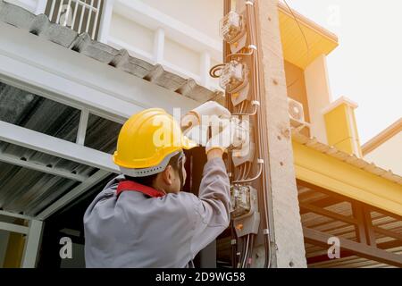 Un électricien en uniforme gris porte des gants et un casque pour installer un compteur de puissance sur un poteau électrique. Banque D'Images