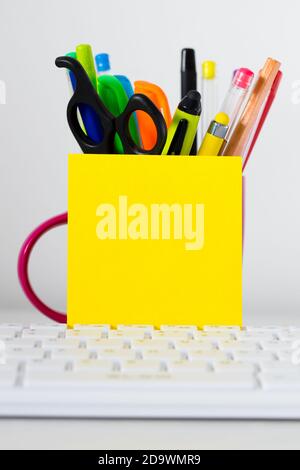 Clavier d'ordinateur, UNE carte vierge jaune avec espace pour le texte, appuyé contre une tasse pleine de stylos, stylos-feutres, crayons et ciseaux. Banque D'Images