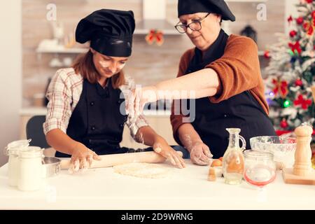 Grand-mère portant un tablier le jour de noël étalant la farine joyeuse joyeuse joyeuse adolescente aidant la femme âgée à préparer des biscuits sucrés pour célébrer les vacances d'hiver. Banque D'Images
