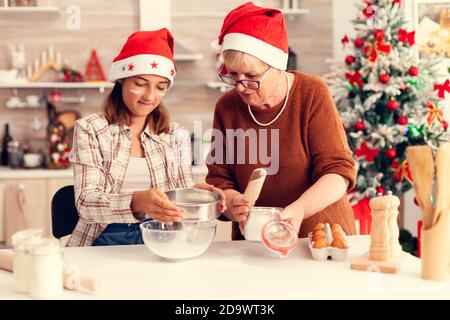 Une famille de plusieurs générations qui fait des biscuits ensemble le jour de noël. Joyeux joyeuse adolescente aidant la femme senior à préparer des petits-circuits pour célébrer les vacances d'hiver en portant le chapeau de père noël. Banque D'Images