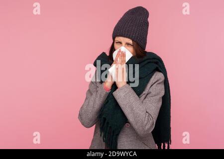 Malade brune femme dans le chapeau et le foulard soufflant son nez dans la serviette, souffrant de nez liquide, allergies saisonnières. Studio d'intérieur isolé sur rose Banque D'Images