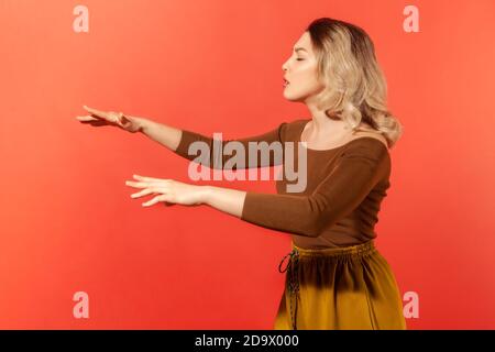 Vue latérale portrait de blonde belle femme en blouse brune debout avec les yeux fermés et les mains étirées, essayant de se déplacer en avant aveugle. St. Intérieure Banque D'Images