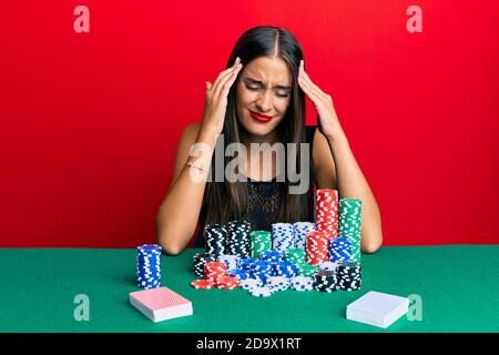 Jeune femme hispanique assis sur la table jouant au poker souffrant de maux de tête désespérés et stressés parce que la douleur et la migraine. Mains sur la tête. Banque D'Images