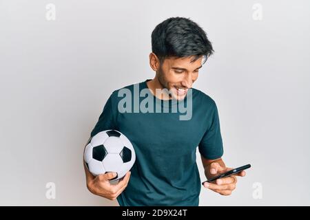 Jeune homme beau tenant le ballon de football regardant l'application de pari de smartphone souriant et riant fort parce que drôle de blague fou. Banque D'Images