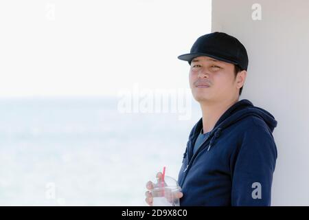 Homme asiatique portant une chemise bleue à manches longues et portant un chapeau tenant un verre en plastique d'eau par le océan Banque D'Images